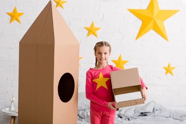 Lächelndes Kind mit Zöpfen, Helm in der Hand und neben Papprakete stehend — Stockfoto