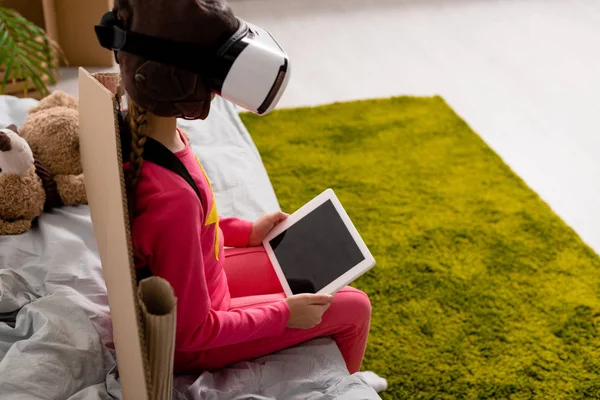 Bambino in auricolare vr seduto sul letto e con in mano tablet digitale con schermo bianco — Foto stock