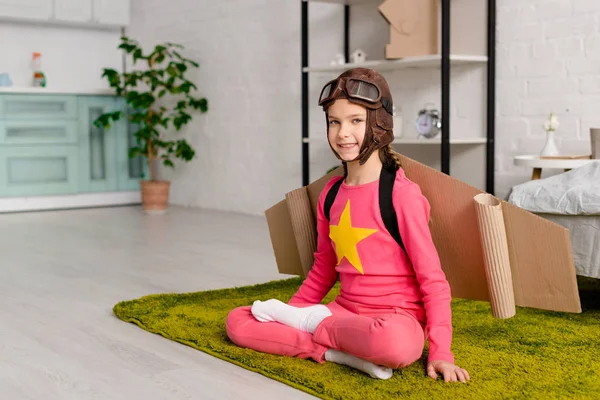 Enfant souriant avec des ailes en carton assis dans la pose du lotus sur le tapis vert — Photo de stock