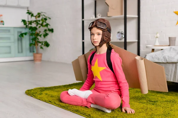 Serious child with cardboard wings sitting in lotus pose on carpet — Stock Photo