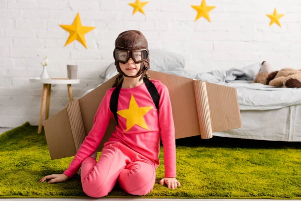 Niño curioso con gafas y casco de vuelo sentado en la alfombra verde - foto de stock