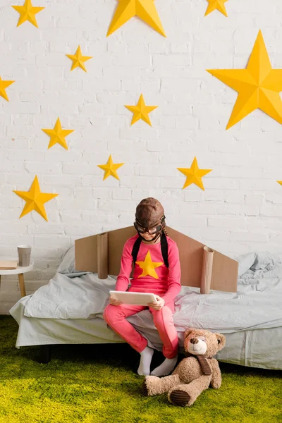 Child in flight helmet sitting on bed and using digital tablet — Stock Photo