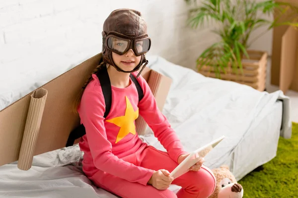 Цікава дитина в льотному шоломі сидить на ліжку і тримає цифровий планшет — стокове фото