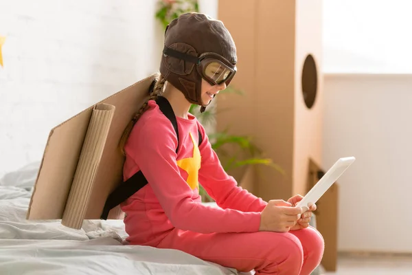 Criança adorável com asas de papelão sentado na cama e usando tablet digital — Fotografia de Stock
