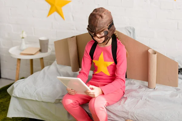 Kid in pink clothes and flight helmet using digital tablet while sitting on bed — Stock Photo