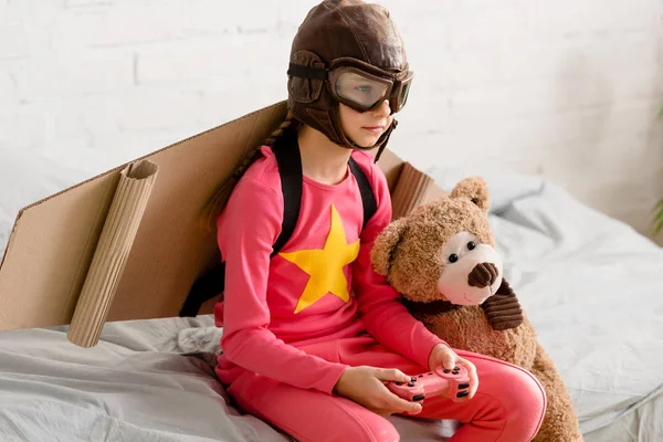 Enfant avec un ours en peluche assis sur le lit et tenant un joystick — Photo de stock