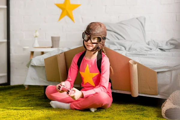 Joyful kid in flight helmet sitting on carpet and playing video game with gamepad — Stock Photo