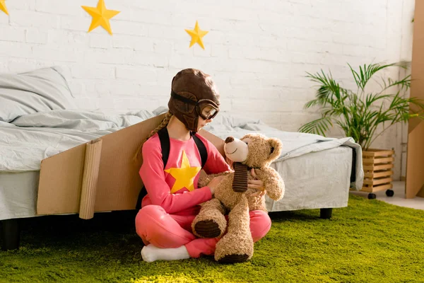 Child with cardboard wings sitting on carpet and looking at teddy bear — Stock Photo