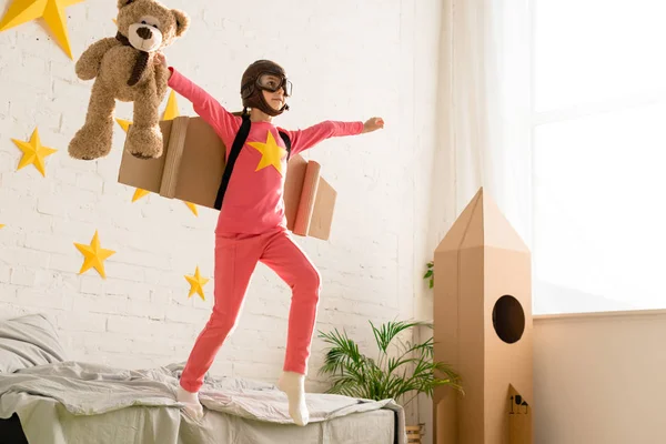 Active child with cardboard wings posing on bed with teddy bear — Stock Photo