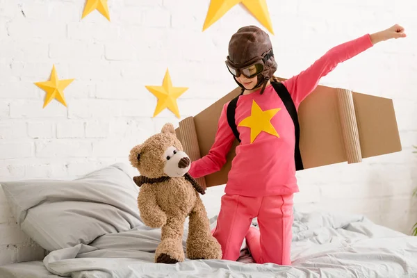 Niño con alas de cartón de pie en la cama y sosteniendo oso de peluche - foto de stock