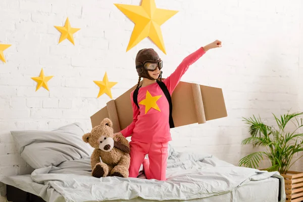 Lächelndes Kind mit Teddybär im Bett und erhobener Hand — Stockfoto