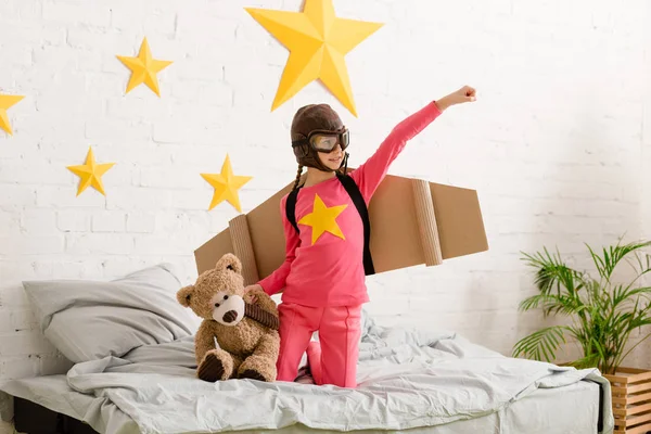 Smiling child with teddy bear standing on bed and holding hand up — Stock Photo