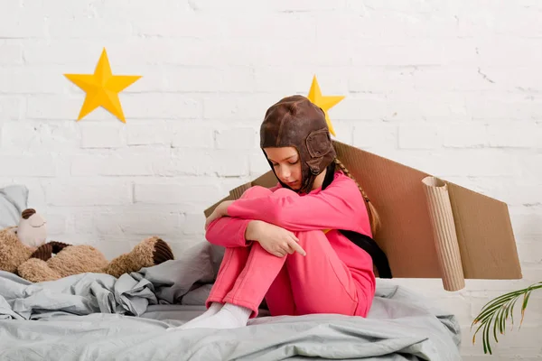 Triste enfant avec des ailes en carton assis sur le lit et embrassant les genoux — Photo de stock