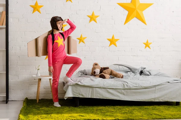 Aufgeregtes Kind mit Pappflügeln steht im Schlafzimmer und schaut weg — Stockfoto