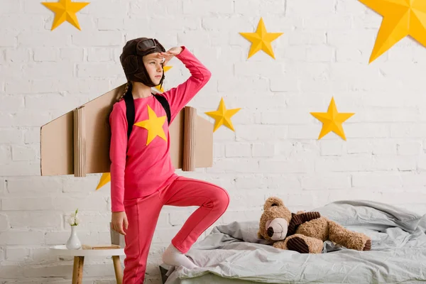 Selbstbewusstes Kind mit Pappflügeln schaut im Schlafzimmer weg — Stockfoto