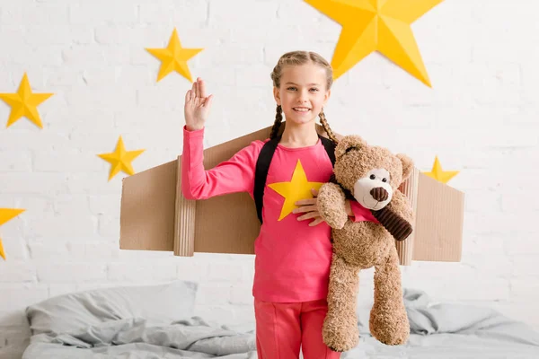 Kind mit Zöpfen hält Teddybär in der Hand und winkt im Schlafzimmer — Stockfoto