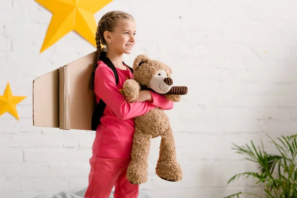 Happy kid with cardboard wings holding teddy bear and looking away — Stock Photo