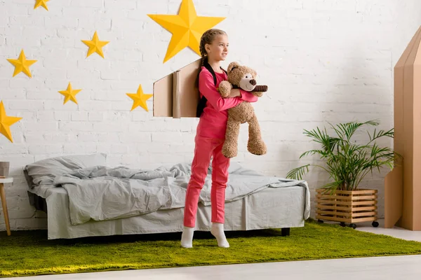 Niño con oso de peluche de pie de puntillas y mirando hacia otro lado en el dormitorio - foto de stock