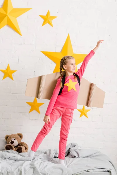 Enfant confiant avec des ailes en carton debout sur le lit avec la main en l'air — Photo de stock