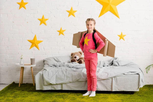 Уверенный ребенок с картонными крыльями стоит с руками акимбо в спальне — стоковое фото