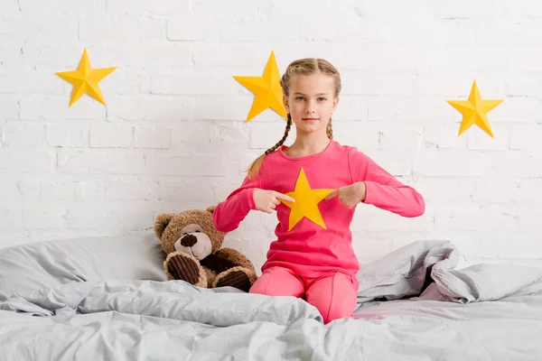 Child with braids sitting on bed and pointing with fingers at yellow star — Stock Photo