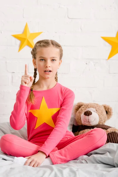 Joli gosse avec un ours en peluche assis sur le lit et tenant le doigt vers le haut — Photo de stock