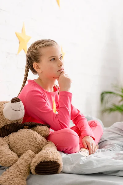 Niedliches, nachdenkliches Kind mit Teddybär auf dem Bett sitzend und Kinn berührend — Stockfoto