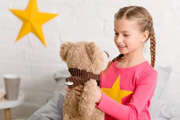 Glückseliges Kind mit Zöpfen blickt lächelnd auf Teddybär — Stockfoto