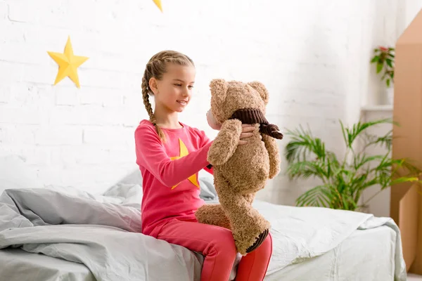 Criança encantadora com tranças sentadas na cama e olhando para ursinho de pelúcia — Fotografia de Stock