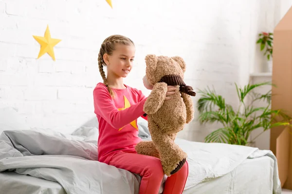 Encantador niño con trenzas sentado en la cama y mirando oso de peluche - foto de stock