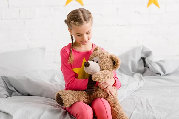 Criança adorável sentado na cama e segurando ursinho de pelúcia nos joelhos — Fotografia de Stock