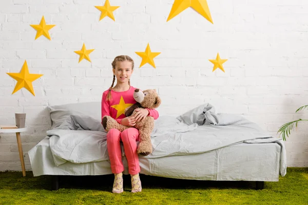 Miúdo rindo sentado na cama com ursinho de pelúcia — Fotografia de Stock