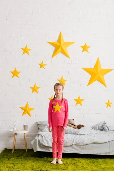 Cheerful kid in pink clothes standing in front of bed — Stock Photo