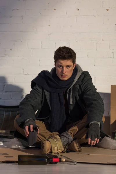 Homeless man sitting on cardboard by white brick wall and holding paper cup — Stock Photo