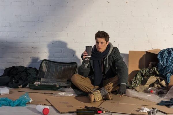 Бездомный держит бумажную чашку, сидя на картонке в окружении мусора. — стоковое фото