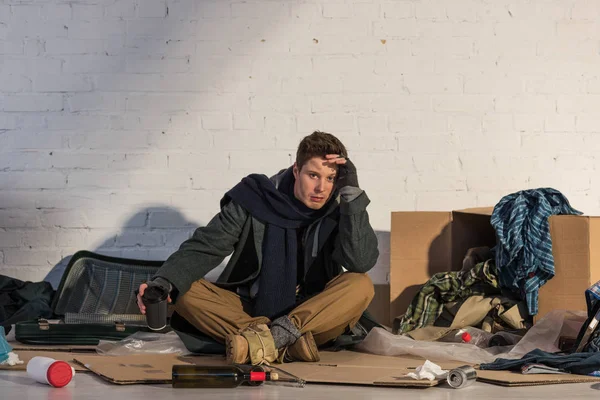 Depressiver Obdachloser mit Pappbecher auf Pappe sitzend, umgeben von Müll — Stockfoto