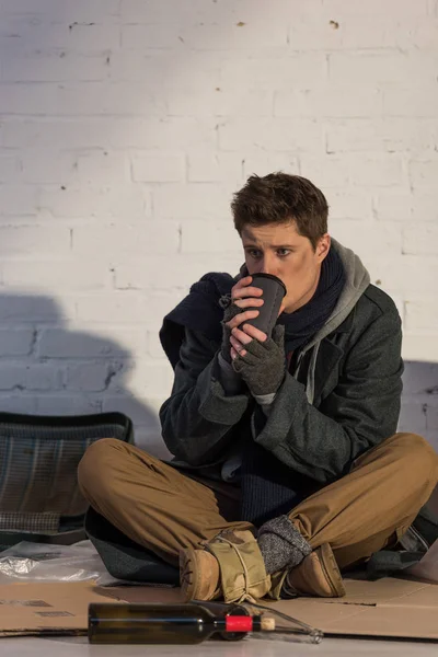 Sad homeless man drinking from paper cup while sitting on cardboard by brick wall — Stock Photo