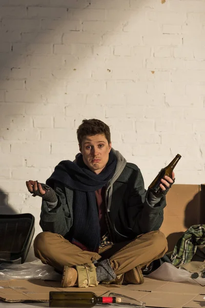 Upset homeless man sitting on cardboard surrounded by rubbish and holding empty alcohol bottle — Stock Photo