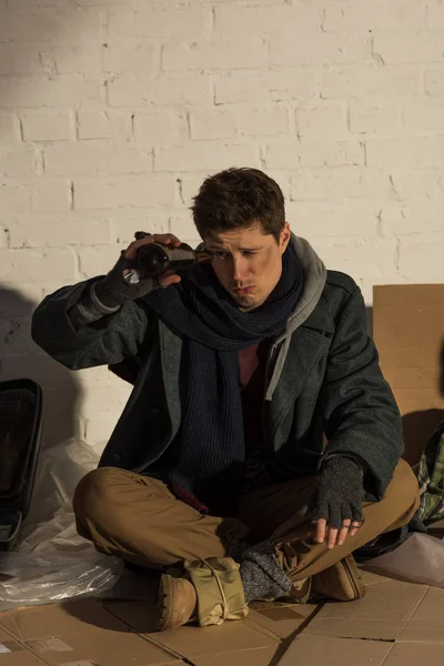 Homeless man looking into empty bottle while sitting on cardboard by brick wall — Stock Photo