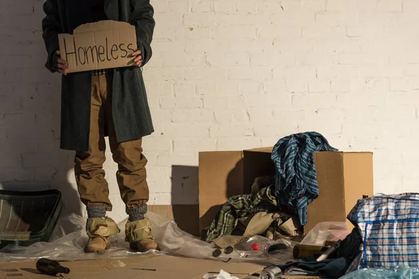 Teilansicht eines Obdachlosen, der auf einer Müllhalde steht und ein Stück Pappe mit der handgeschriebenen Aufschrift 