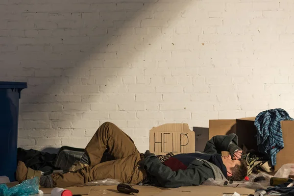Бездомный лежит на картонке в окружении мусора под белой кирпичной стеной — стоковое фото