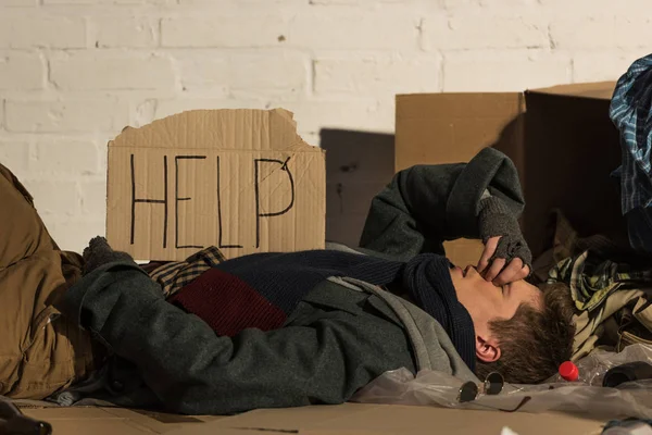 Бездомный несчастный лежит на картонке, с надписью 