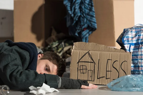 Депрессивный бездомный лежит на картонке в мусорной свалке, с символом дома и надписью 