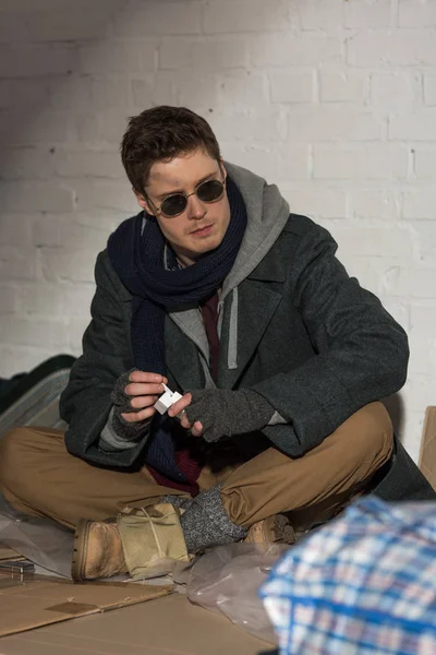 Homeless man with cigarette pack sitting on cardboard by brick wall — Stock Photo