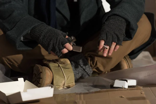 Vista parcial del hombre sin hogar con guantes sin dedos sosteniendo encendedor y cigarrillo - foto de stock