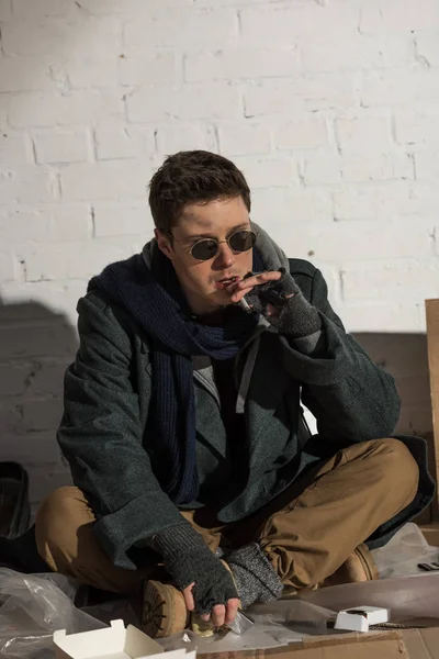 Homeless man smoking cigarette while sitting by white brick wall — Stock Photo