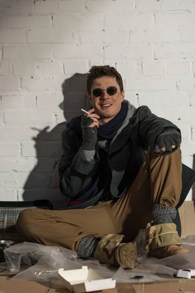 Sonriente hombre sin hogar fumando mientras está sentado junto a la pared de ladrillo - foto de stock