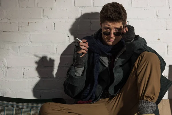 Homeless man in dark glasses smoking while sitting by white brick wall — Stock Photo