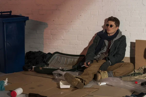 Бездомный сидит на помойке у кирпичной стены и курит сигарету. — стоковое фото