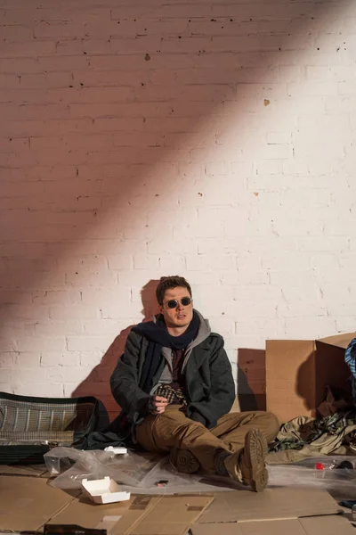 Homeless man surrounded by rubbish sitting by white brick wall — Stock Photo